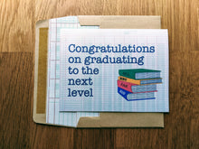 Load image into Gallery viewer, Accounting greeting card, with images of four stacked CPA exam review books titled &quot;REG&quot;, &quot;BEC&quot;, &quot;FAR&quot;, and &quot;AUD&quot;, with text that reads &quot;Congratulations on graduating to the next level&quot;, atop an envelope lined in ledger paper 

