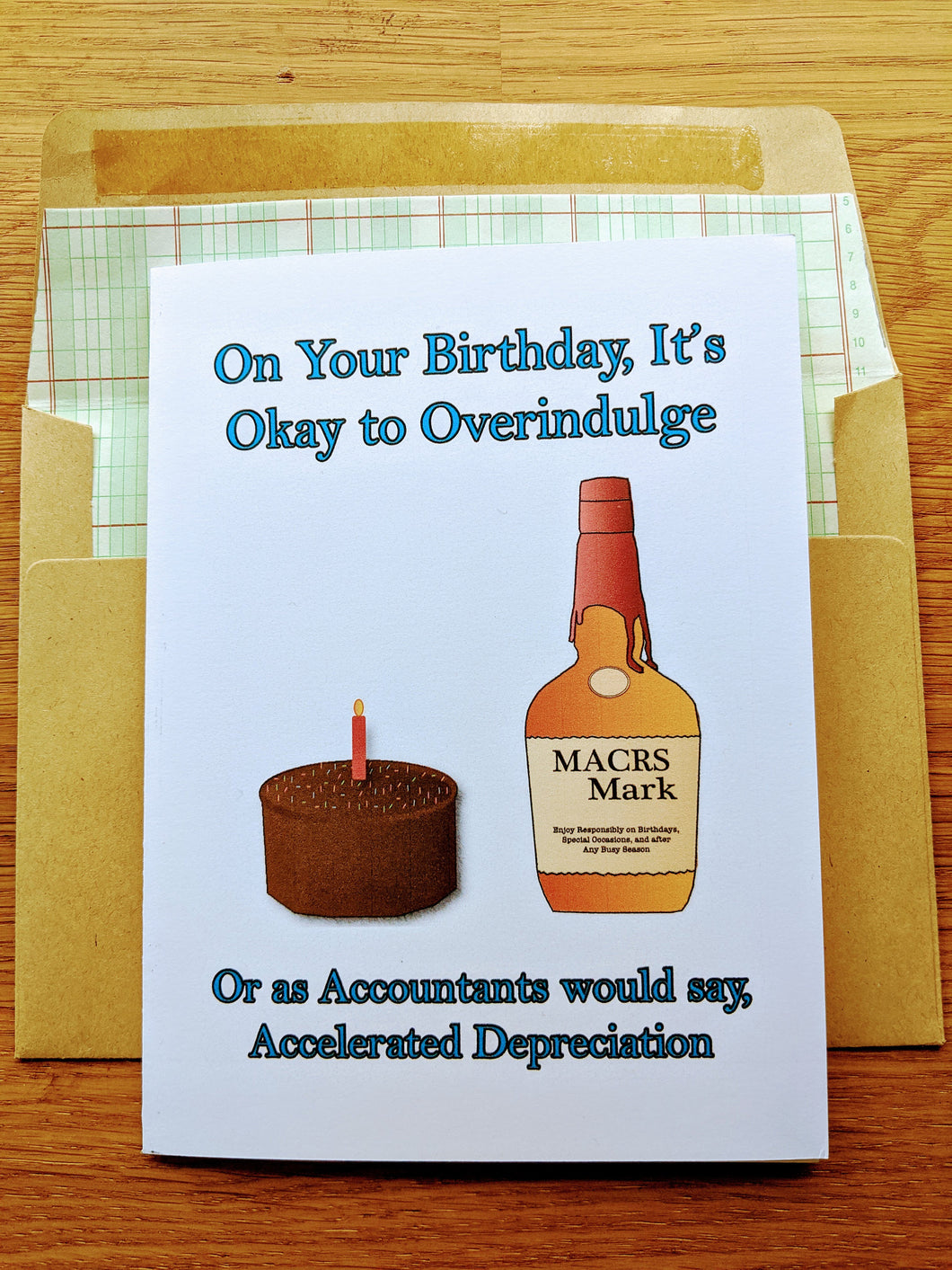 Accounting greeting card, with a birthday cake and a bottle labeled MACRS Mark that parodies a bottle of Makers Mark, atop an envelope lined in ledger paper